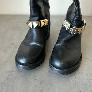 Peut inclure: Paire de bottes en cuir noir avec une bride de cheville clout&eacute;e dor&eacute;e. Les bottes ont un bout arrondi et un petit talon. Elles sont pr&eacute;sent&eacute;es sur une surface grise.