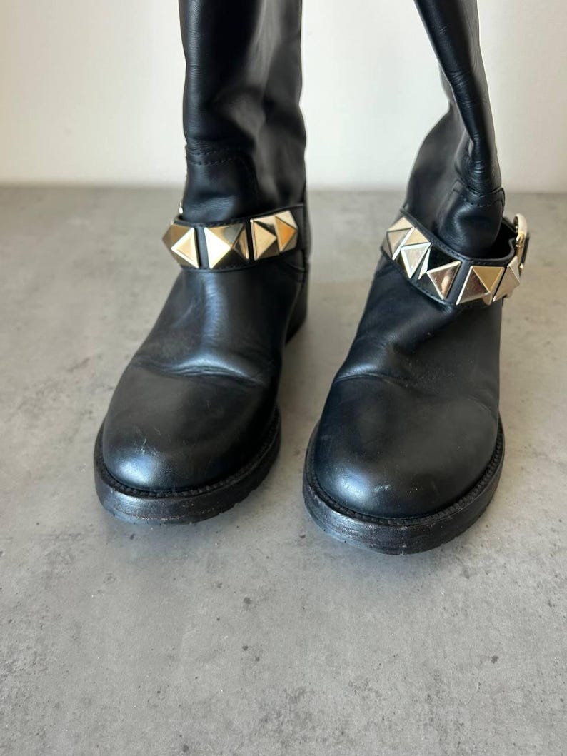 Peut inclure: Paire de bottes en cuir noir avec une bride de cheville clout&eacute;e dor&eacute;e. Les bottes ont un bout arrondi et un petit talon. Elles sont pr&eacute;sent&eacute;es sur une surface grise.