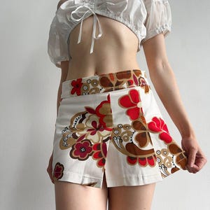 May include: A white crop top with puff sleeves and a tie front, worn with a white mini skirt. The skirt has a floral pattern in red, brown, and tan. The outfit is a retro style.