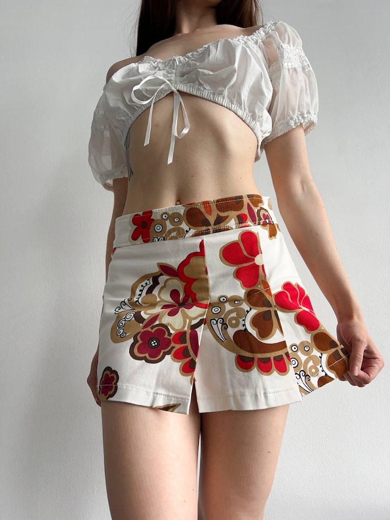 May include: A white crop top with puff sleeves and a tie front, worn with a white mini skirt. The skirt has a floral pattern in red, brown, and tan. The outfit is a retro style.