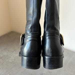 Peut inclure: Paire de bottes d'&eacute;quitation en cuir noir avec boucles dor&eacute;es. Les bottes ont un talon de hauteur moyenne et une finition lisse et polie. L'image montre l'arri&egrave;re des bottes, soulignant les d&eacute;tails du cuir et de la quincaillerie.