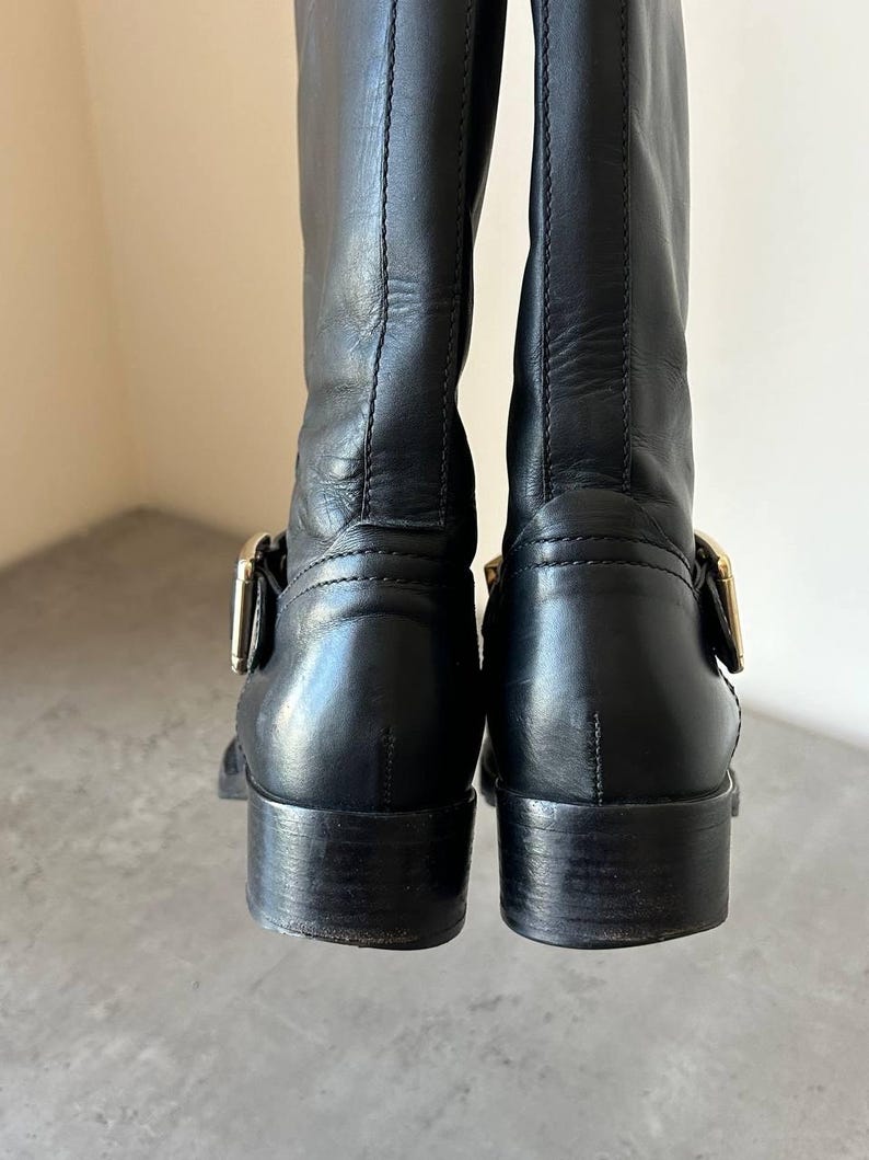 Peut inclure: Paire de bottes d'&eacute;quitation en cuir noir avec boucles dor&eacute;es. Les bottes ont un talon de hauteur moyenne et une finition lisse et polie. L'image montre l'arri&egrave;re des bottes, soulignant les d&eacute;tails du cuir et de la quincaillerie.
