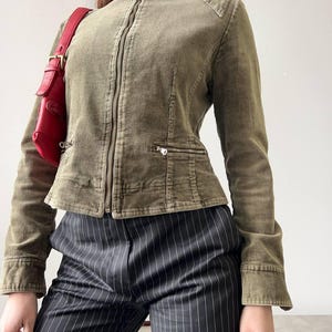 May include: An olive green corduroy jacket with a full zip, stand-up collar, and two small pockets. The jacket is paired with black and white pinstripe trousers. A red bag is also visible.
