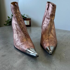 May include: Rose gold ankle boots with silver metal pointed toes. The boots have a textured finish and a high heel. The boots are displayed on a grey surface.