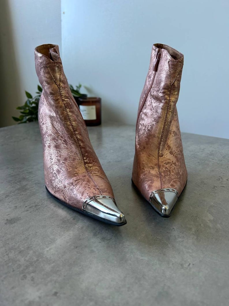 May include: Rose gold ankle boots with silver metal pointed toes. The boots have a textured finish and a high heel. The boots are displayed on a grey surface.