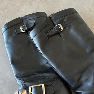 Peut inclure: Paire de bottes d'&eacute;quitation en cuir noir avec boucles et clous dor&eacute;s. Les bottes ont un design classique avec une hauteur mi-mollet et une finition lisse et polie. Les boucles sont rectangulaires et les clous en forme de pyramide.