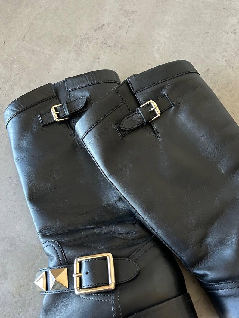 Peut inclure: Paire de bottes d'&eacute;quitation en cuir noir avec boucles et clous dor&eacute;s. Les bottes ont un design classique avec une hauteur mi-mollet et une finition lisse et polie. Les boucles sont rectangulaires et les clous en forme de pyramide.