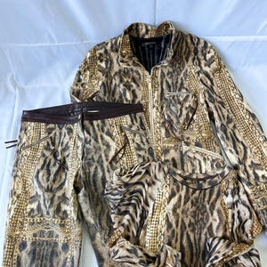 May include: A matching set consisting of a long-sleeve zip-up jacket, trousers, and a long-sleeve top, all featuring a brown and black animal print pattern with gold-tone chain link accents. The trousers have a dark brown waistband and zipper pockets.