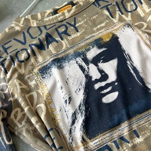 May include: A tan long-sleeved shirt with the word "REVOLUTIONARY" in dark blue lettering. The shirt features a graphic of a person's face in a rectangular frame. The shirt is from Urban Outfitters.