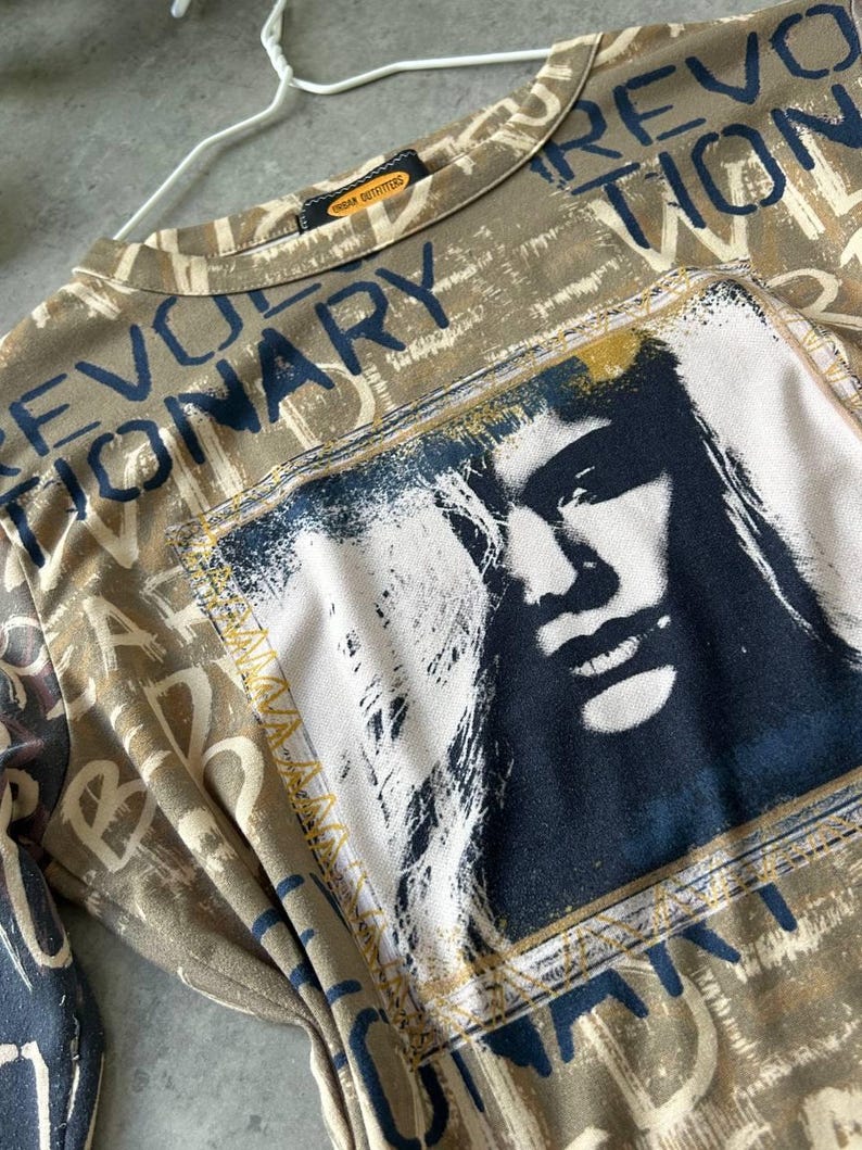 May include: A tan long-sleeved shirt with the word "REVOLUTIONARY" in dark blue lettering. The shirt features a graphic of a person's face in a rectangular frame. The shirt is from Urban Outfitters.