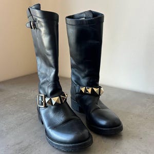Peut inclure: Paire de bottes noires en cuir, montant jusqu'aux genoux. Chaque botte est orn&eacute;e d'une lani&egrave;re clout&eacute;e dor&eacute;e autour de la cheville et d'une boucle. Les bottes ont un bout arrondi et une semelle &eacute;paisse.