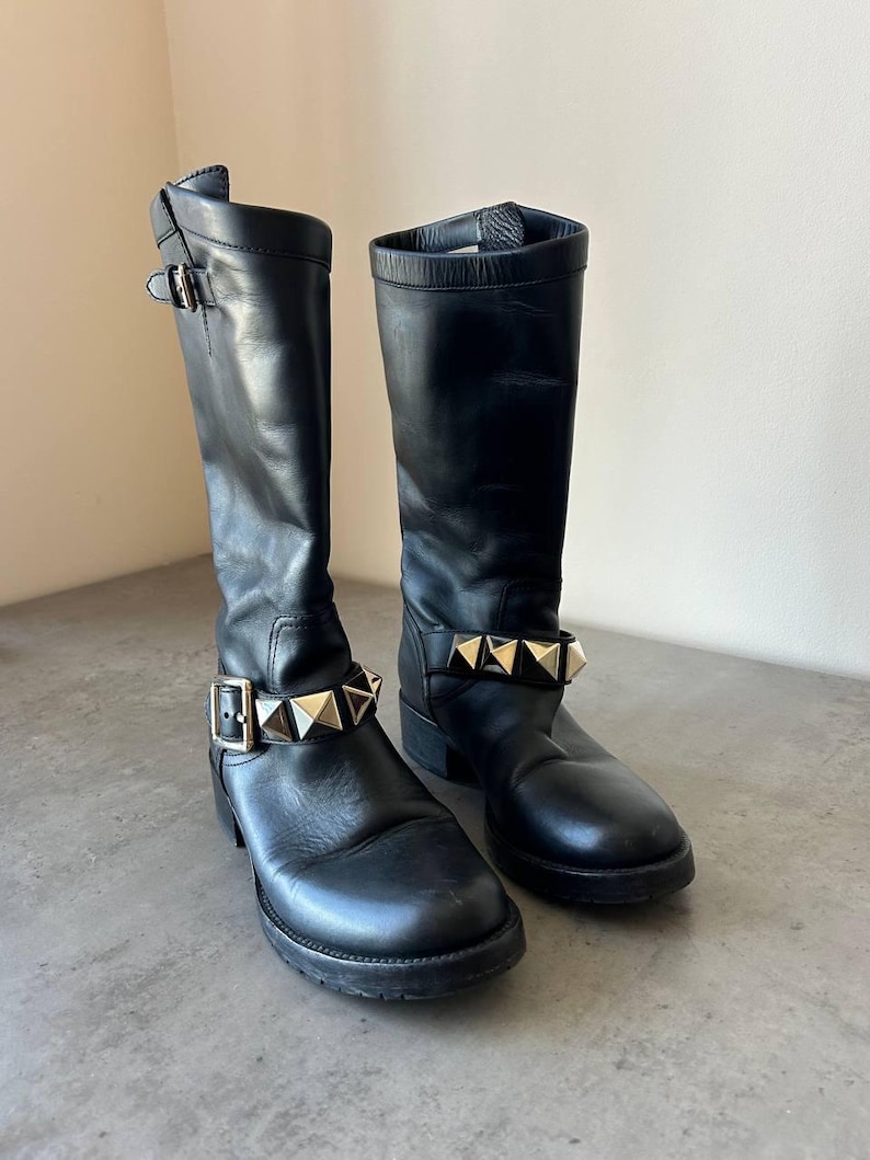 Peut inclure: Paire de bottes noires en cuir, montant jusqu'aux genoux. Chaque botte est orn&eacute;e d'une lani&egrave;re clout&eacute;e dor&eacute;e autour de la cheville et d'une boucle. Les bottes ont un bout arrondi et une semelle &eacute;paisse.