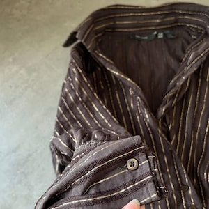 May include: A brown button-down shirt with vertical gold stripes. The long-sleeved shirt has a classic collar and buttoned cuffs. The fabric appears to have a subtle sheen, and the buttons are a dark, metallic color.