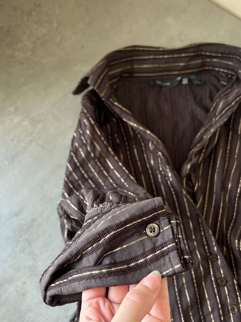 May include: A brown button-down shirt with vertical gold stripes. The long-sleeved shirt has a classic collar and buttoned cuffs. The fabric appears to have a subtle sheen, and the buttons are a dark, metallic color.