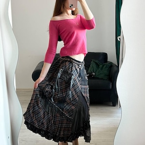May include: A hot pink off-the-shoulder sweater with three-quarter sleeves. The outfit includes a long, layered skirt with a plaid pattern in shades of gray, black, and red. The skirt has a ruffled hem. Black heeled sandals complete the look.