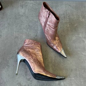 May include: A pair of rose gold ankle boots with silver detailing. These boots feature pointed toes with silver tips, high stiletto heels, and a side zip. The metallic finish gives them a glamorous appearance.