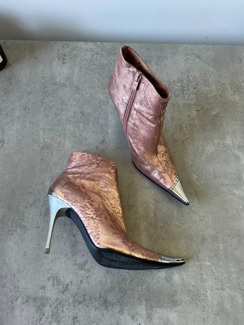 May include: A pair of rose gold ankle boots with silver detailing. These boots feature pointed toes with silver tips, high stiletto heels, and a side zip. The metallic finish gives them a glamorous appearance.