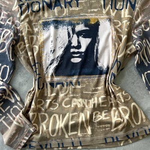 May include: A long-sleeved shirt with a brown and beige base color, featuring a graphic of a woman's face and the words "REVOLUTIONARY" and "BROKEN HEARTS" in a distressed font. The shirt has a crew neck and a tag that says "URBAN OUTFITTERS".