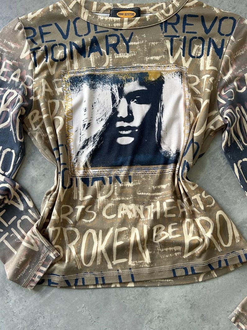 May include: A long-sleeved shirt with a brown and beige base color, featuring a graphic of a woman's face and the words "REVOLUTIONARY" and "BROKEN HEARTS" in a distressed font. The shirt has a crew neck and a tag that says "URBAN OUTFITTERS".