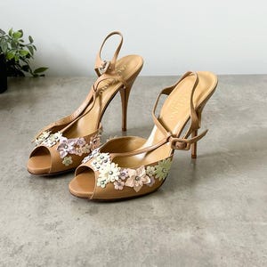 May include: A pair of tan high-heeled sandals with floral embellishments. The open-toe shoes have thin ankle straps and stiletto heels. The flowers are in shades of pink, white, and green. The sandals are displayed on a gray surface.