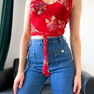 May include: A sleeveless red crop top featuring a v-neckline and a floral pattern in shades of purple, orange, and green, with a tie-front detail at the waist, paired with high-waisted blue denim jeans that have two front pockets and silver buttons.
