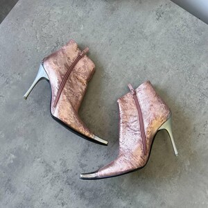May include: Rose gold ankle boots with silver detailing. The boots feature a pointed toe, a zip fastening, and a stiletto heel. The heels are approximately 10 cm high.