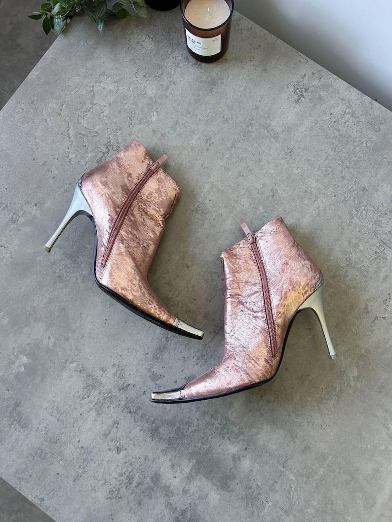 May include: Rose gold ankle boots with silver detailing. The boots feature a pointed toe, a zip fastening, and a stiletto heel. The heels are approximately 10 cm high.