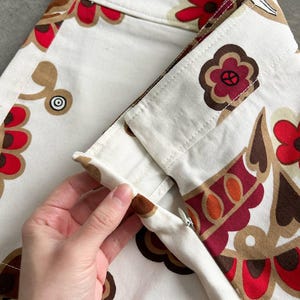 May include: Close-up of a white skirt with a colourful floral pattern in red, brown, and tan. The skirt has a zip and is being held open, showcasing the interior construction. The design features stylised flowers and geometric shapes.