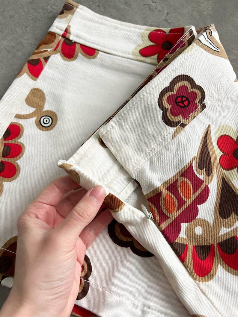 May include: Close-up of a white skirt with a colourful floral pattern in red, brown, and tan. The skirt has a zip and is being held open, showcasing the interior construction. The design features stylised flowers and geometric shapes.