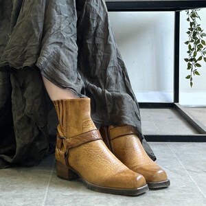 May include: A pair of tan leather ankle boots with a textured finish. The boots feature a strap detail across the ankle and a small block heel. The boots are worn with a long, flowing, dark green skirt.
