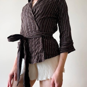 May include: Brown wrap top with vertical white pinstripes, a wide collar, and a matching tie belt. The three-quarter sleeves and tailored fit create a sophisticated look. Paired with white pleated shorts, this top offers a stylish ensemble.