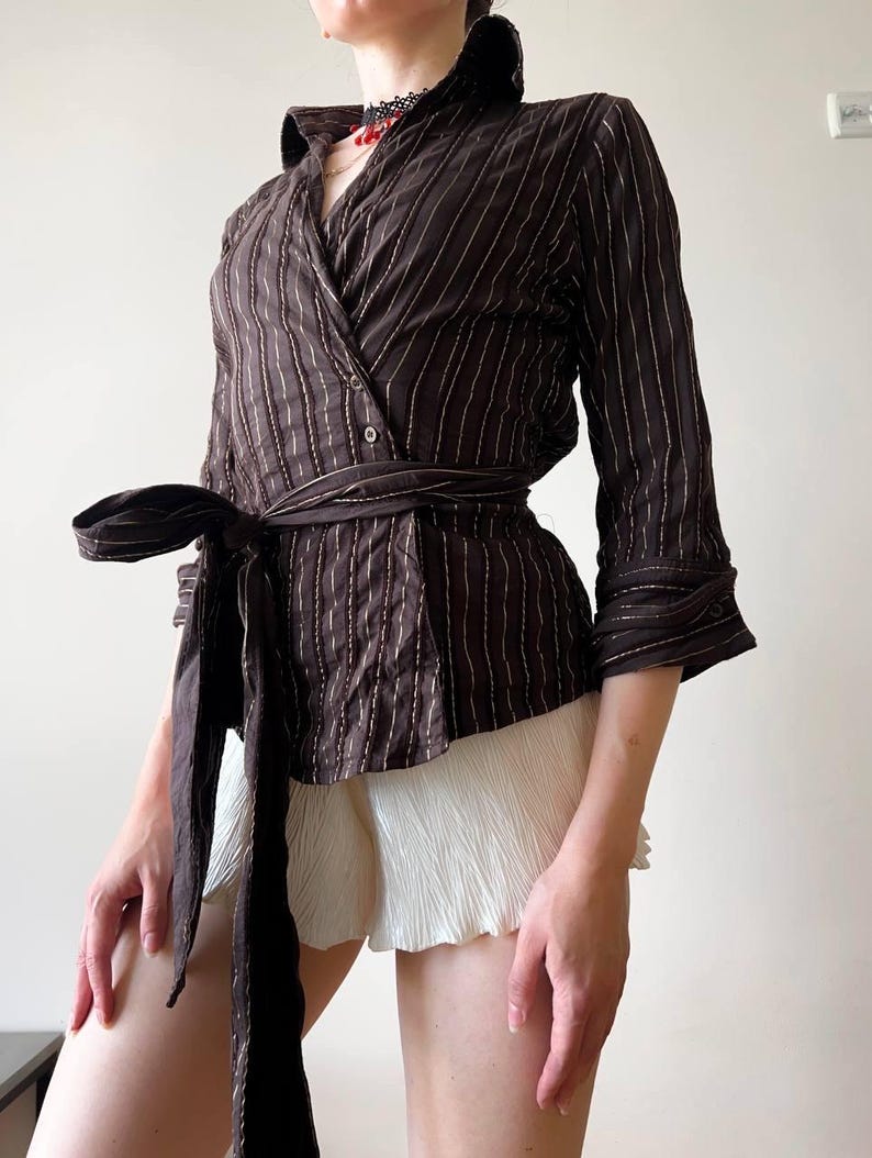 May include: Brown wrap top with vertical white pinstripes, a wide collar, and a matching tie belt. The three-quarter sleeves and tailored fit create a sophisticated look. Paired with white pleated shorts, this top offers a stylish ensemble.