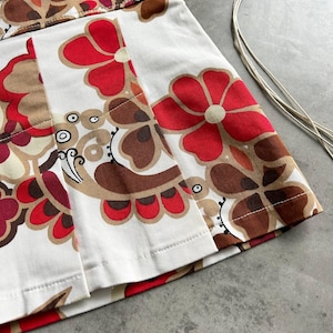 May include: A white pleated skirt with a striking floral design in red, brown, and beige. The skirt has a high waist and a classic A-line shape. The floral pattern is bold and retro, with large stylized flowers.