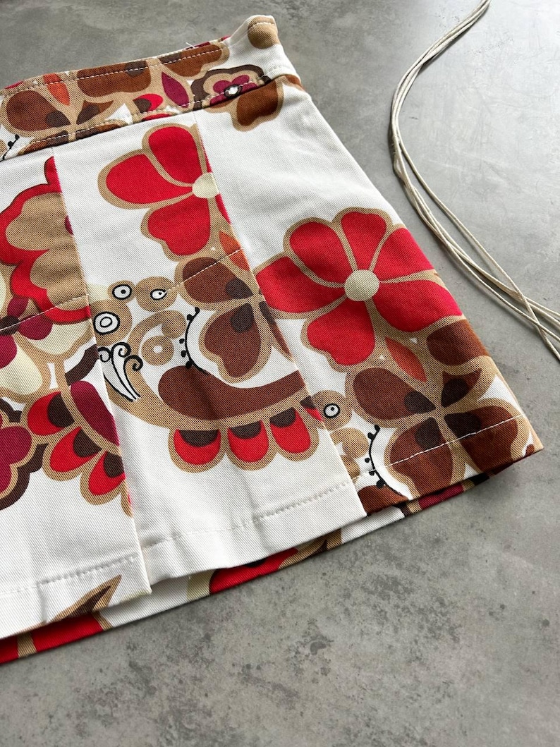 May include: A white pleated skirt with a striking floral design in red, brown, and beige. The skirt has a high waist and a classic A-line shape. The floral pattern is bold and retro, with large stylized flowers.