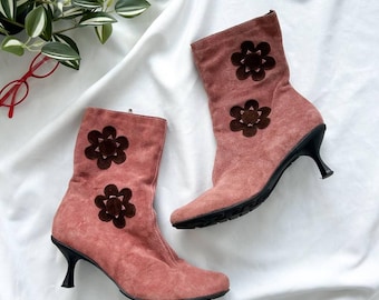 May include: A pair of pink suede ankle boots with dark brown flower appliqués. The boots have a pointed toe and a black, curved heel. The boots are set against a white background.