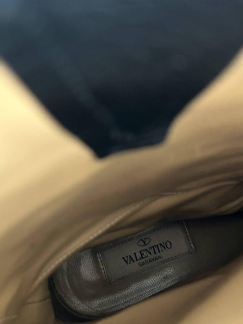 Peut inclure: Vue int&eacute;rieure d'une botte en cuir beige. La semelle int&eacute;rieure porte une &eacute;tiquette rectangulaire avec le logo "Valentino Garavani" en noir. L'int&eacute;rieur de la botte est beige clair, avec une ombre sombre en haut.