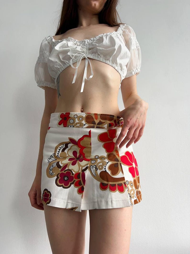 May include: A white crop top with short, sheer sleeves and a tie front, worn with a mini skirt. The skirt has a white base with a repeating floral pattern in red, brown, and tan. The outfit has a retro, 70s style.