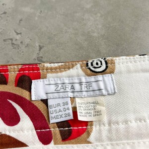 May include: Close-up of white cotton trousers featuring a pattern of red, brown, and black shapes. The label reads "ZARA TRF" and includes size information. The trousers are made in Spain.