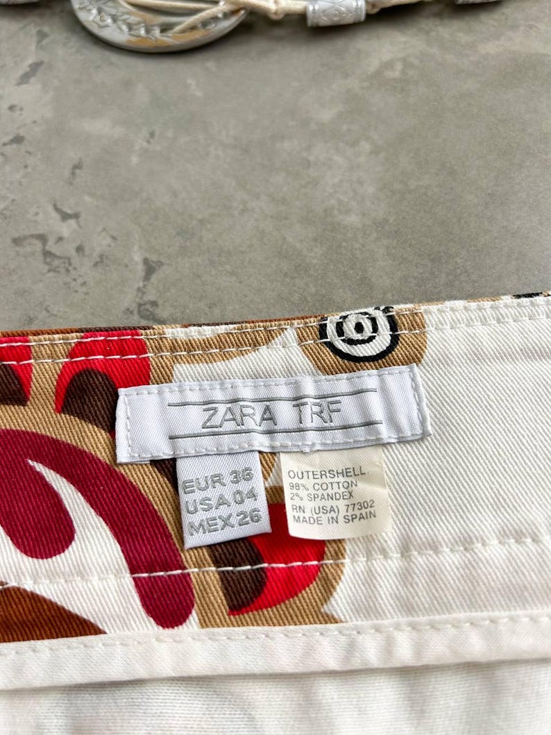 May include: Close-up of white cotton trousers featuring a pattern of red, brown, and black shapes. The label reads "ZARA TRF" and includes size information. The trousers are made in Spain.