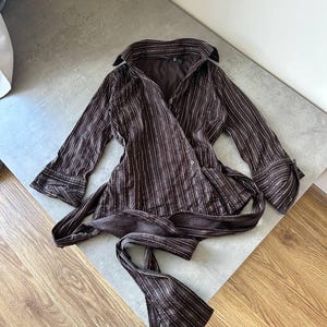 May include: A brown wrap top with vertical white pinstripes. The top has long sleeves with buttoned cuffs and a collar. A matching fabric belt is tied at the waist. The garment is made of a lightweight material.