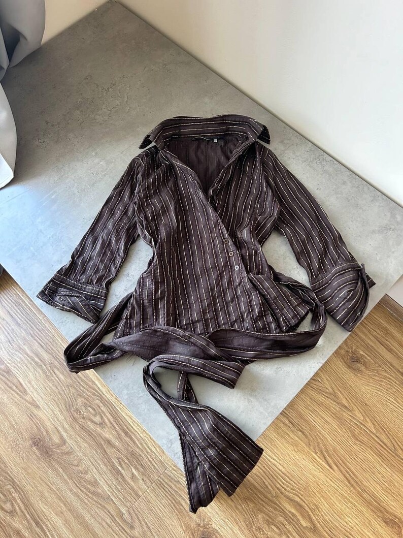 May include: A brown wrap top with vertical white pinstripes. The top has long sleeves with buttoned cuffs and a collar. A matching fabric belt is tied at the waist. The garment is made of a lightweight material.