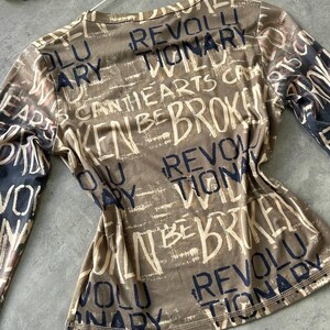 May include: A long-sleeved top in shades of brown and beige, covered in a distressed text design. The words "REVOLUTIONARY", "BROKEN", and "HEARTS" are visible. The top is hanging on a white hanger.