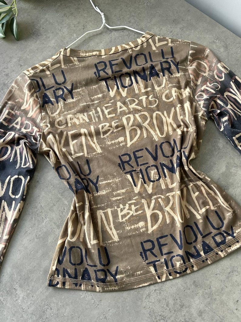 May include: A long-sleeved top in shades of brown and beige, covered in a distressed text design. The words "REVOLUTIONARY", "BROKEN", and "HEARTS" are visible. The top is hanging on a white hanger.