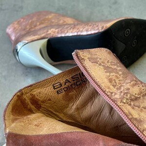 May include: Rose gold ankle boots with a metallic finish. The boots have a pointed toe, a stiletto heel, and a zipper closure. The heel is approximately 10 cm high. The interior of the boot is a light brown colour, and the brand name "BASIC EDITION" is visible.