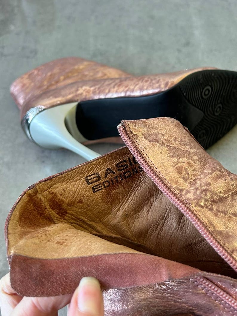 May include: Rose gold ankle boots with a metallic finish. The boots have a pointed toe, a stiletto heel, and a zipper closure. The heel is approximately 10 cm high. The interior of the boot is a light brown colour, and the brand name "BASIC EDITION" is visible.
