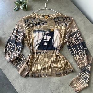 May include: A long-sleeved top with a brown and black color scheme. The shirt features a graphic of a face and the words "REVOLUTIONARY" and "BROKEN" in a distressed font. The top is hanging on a white hanger.