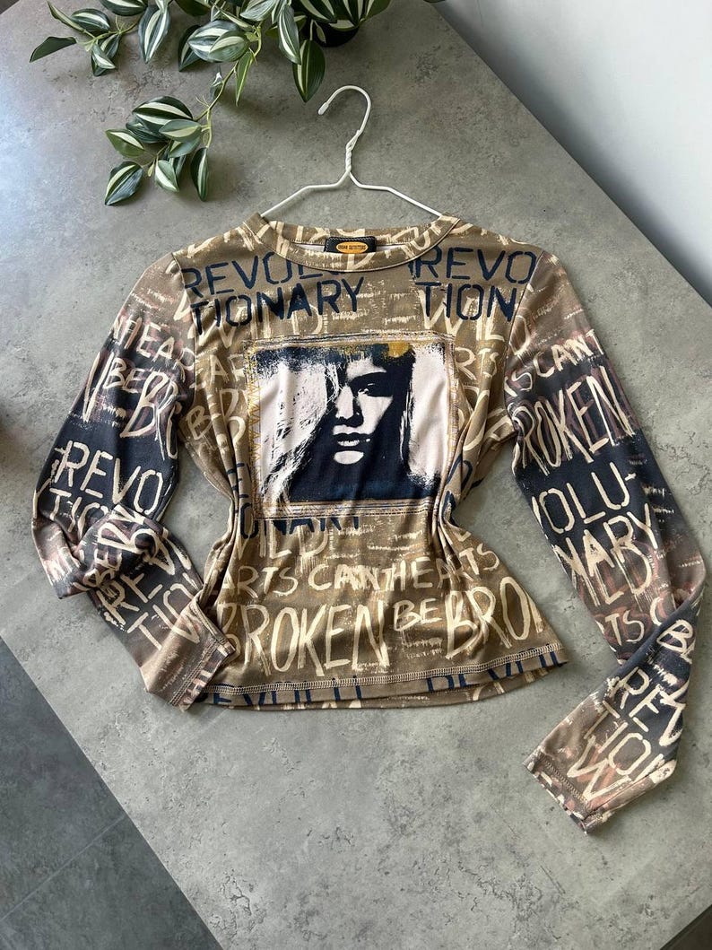 May include: A long-sleeved top with a brown and black color scheme. The shirt features a graphic of a face and the words "REVOLUTIONARY" and "BROKEN" in a distressed font. The top is hanging on a white hanger.