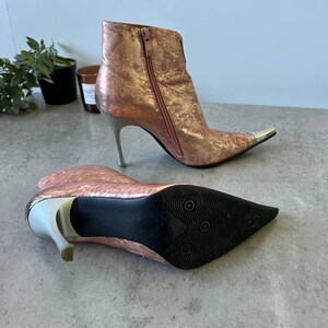 May include: Metallic rose gold ankle boots with pointed toes and stiletto heels. The boots have silver heels and toe caps, and a zip closure. The sole is black.