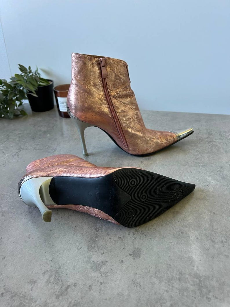 May include: Metallic rose gold ankle boots with pointed toes and stiletto heels. The boots have silver heels and toe caps, and a zip closure. The sole is black.