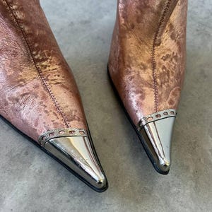 May include: Rose gold ankle boots with pointed toes and silver metal toe caps. The boots have a textured metallic finish and a sleek design. The boots are a stylish footwear choice.